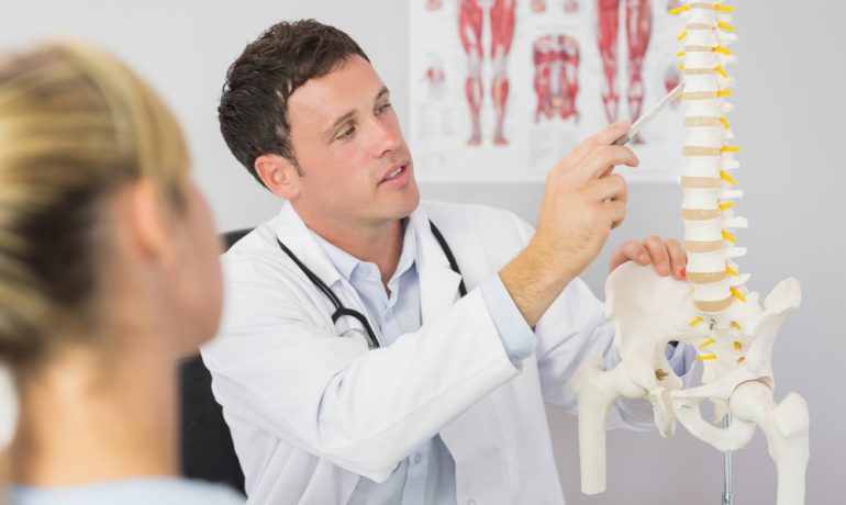 The doctor is sitting in a chair while holding a skeleton model and pointing to the spine with a pen while appearing to be explaining the issue with the spine to the patient.
